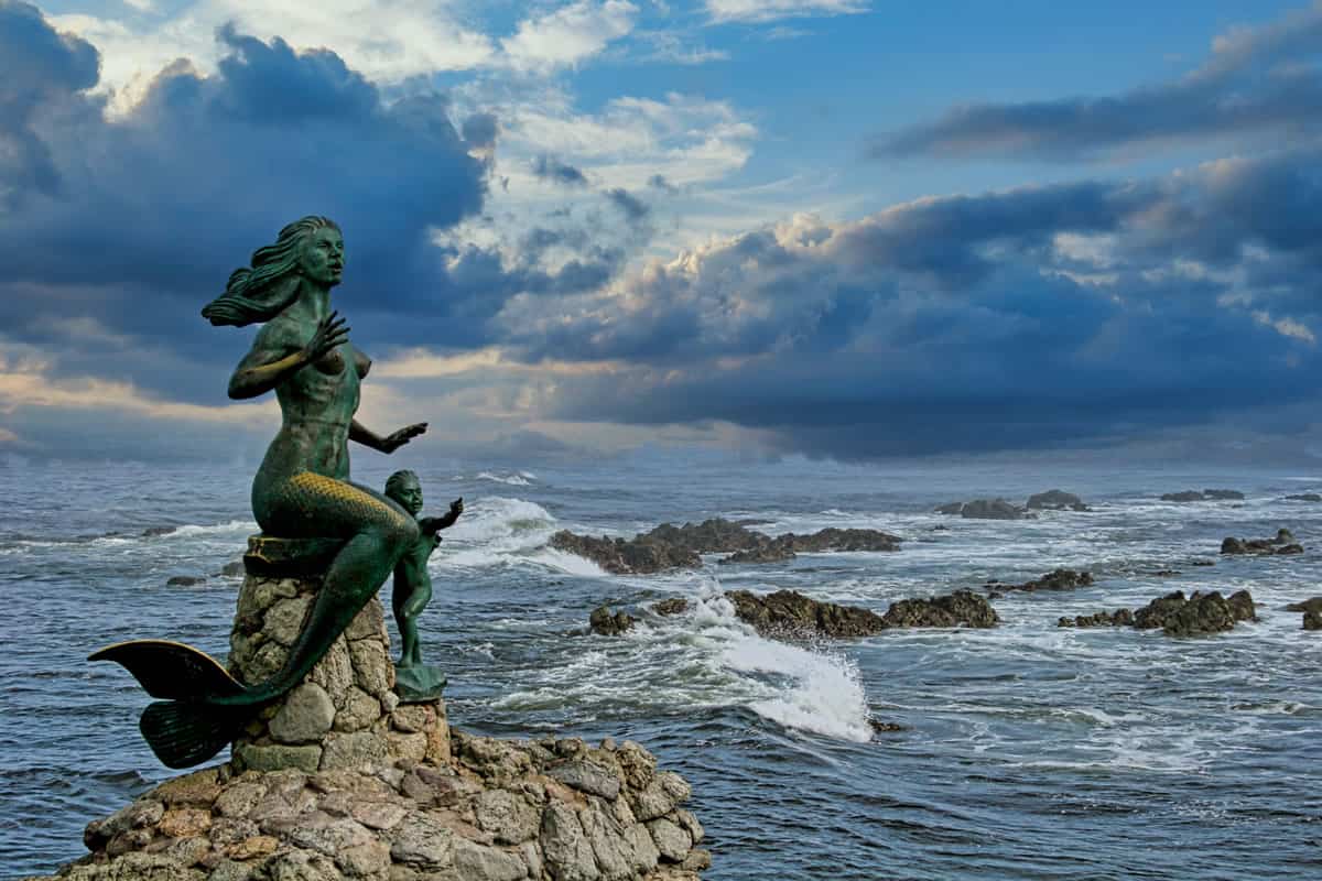 Una estatua de una sirena sentada sobre una roca frente a un cielo tormentoso.