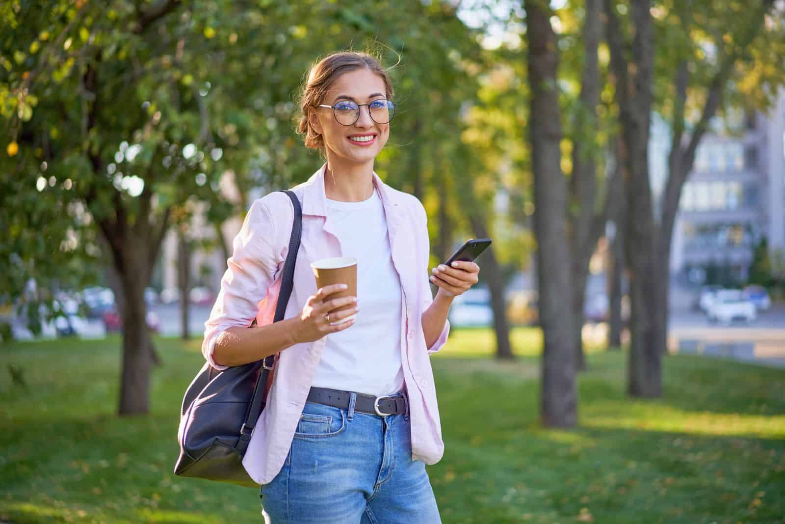 Una mujer joven sosteniendo una taza de café y un teléfono celular en el parque.
