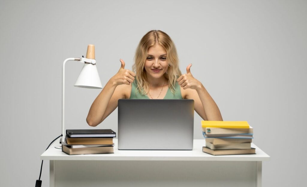 Una mujer joven está sentada en un escritorio con una computadora portátil y levantando el pulgar.